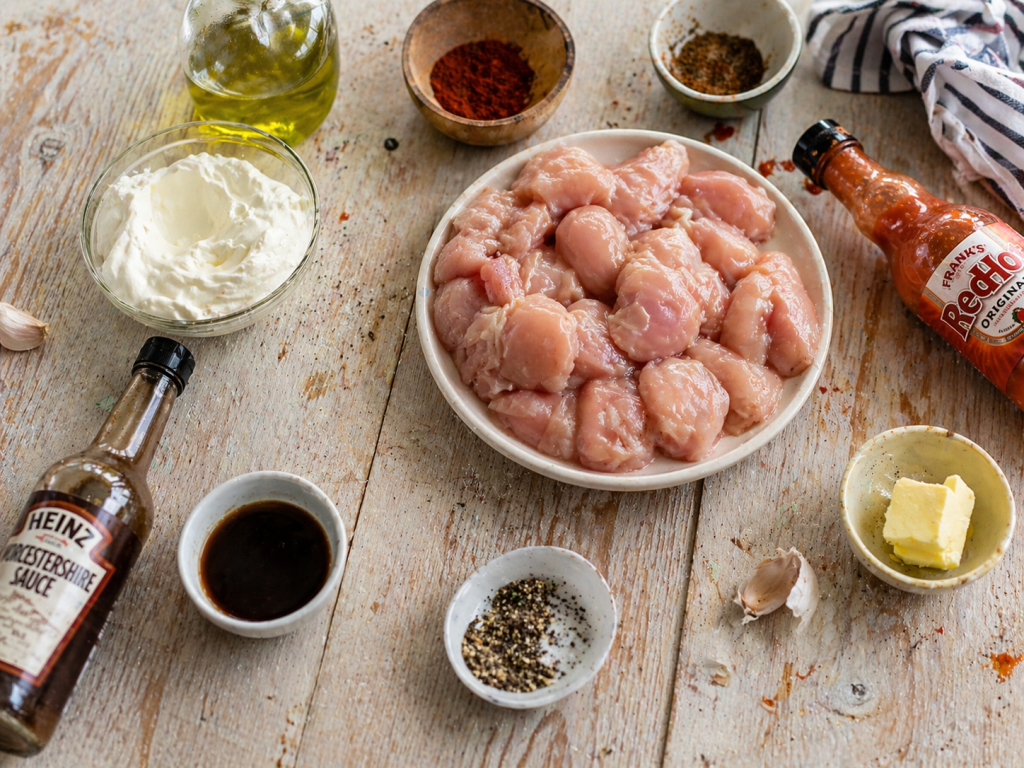 ingredients for buffalo chicken recipe laid out on a wooden table including raw chicken hot sauce butter garlic and spices