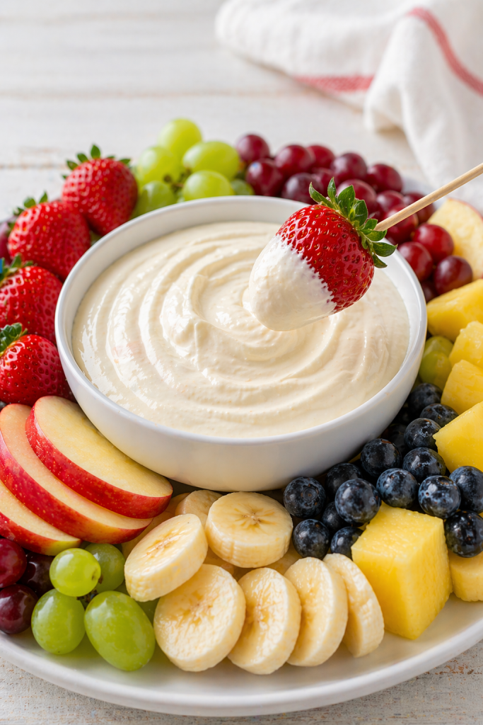 strawberry on a stick dipped in creamy fruit dip with fresh fruit platter in the background close up