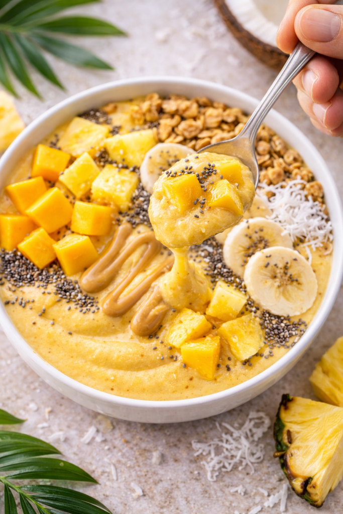 Spoon lifting thick tropical smoothie bowl with mango, banana, chia seeds, and granola
